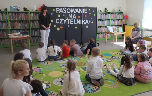 Na zdjęciu znajdują się dzieci siedzące na dywanie, przed nimi stoi kobieta, obok niej znajduje się stolik, a po drugiej stronie czarna tablica z kwiatami i napisem pasowanie na czytelnika. W tle znajdują się półki z książkami.