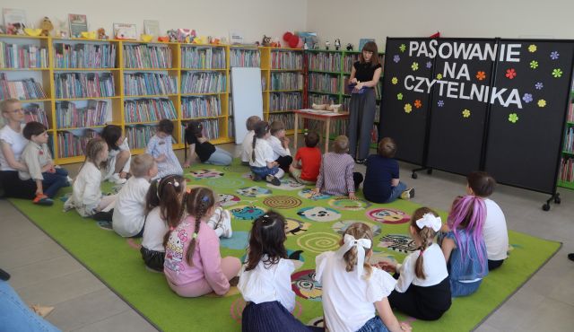 Na zdjęciu znajdują się dziecie na dywanie przed nimi stoi kobieta po lewej stronie znajduje się stolik, a po prawej tablica z kwiatami i napisem pasowanie na czytelnika. W tle znajdują się półki z książkami.