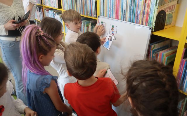 Na zdjęciu znajduje się sześcioro przedszkolaków, którzy przyczepiają rysunki postaci z bajek do tablicy magnetycznej. W tle znajdują się półki z książkami.