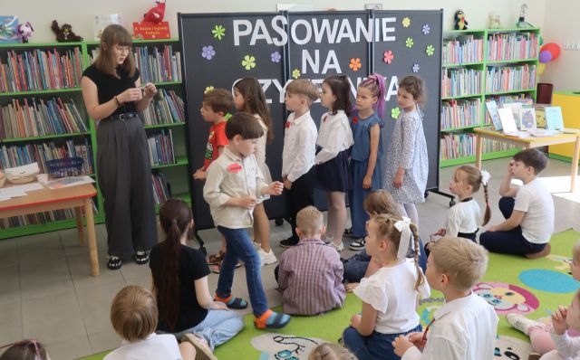 Na zdjęciu znajdują się dzieci na dywanie przed nimi stoi kobieta oraz sześć przedszkolaków ustawionych jeden za drugim. Za nimi znajduje się tablica z napisem, po lewej stronie zdjęcia znajduje się stolik. Jeden chłopiec przechodzi przez dywan. W tle znajdują się półki z książkami.