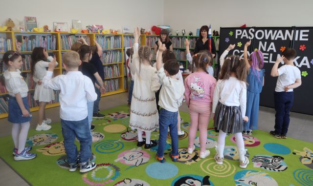 Na zdjęciu znajdują się przedszkolaki na dywanie, które mają podniesione do góry dwa palce. Przed nimi stoją dwie kobiety, jedna z nich ma duży ołówek do pasowania. W tle znajduje się tablica z napisem oraz półki z książkami.