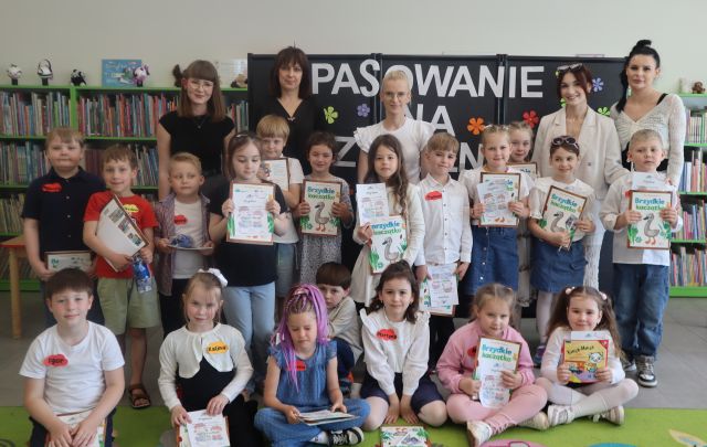 Na zdjęciu znajduje się dziewiętnaście przedszkolaków oraz pięć kobiet i wszyscy wspólnie stoją do zdjęcia. Przedszkolaki trzymają w rękach dyplomy i książki. W tle znajduje się czarna tablica z napisem oraz półki z ksiażkami.