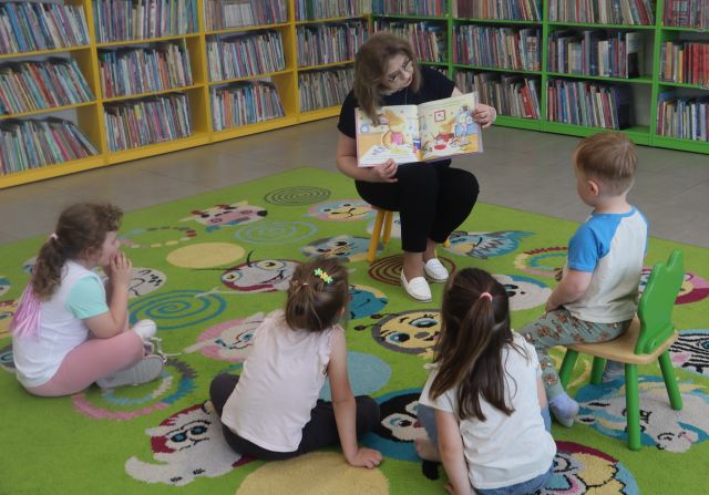 Na zdjęciu znajduje się czwórka dzieci, które siedzą na dywanie przed kobietą, która pokazuje im ilustracje w książce. W tle znajdują się półki z książkami.