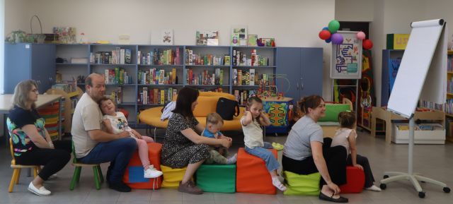 Na zdjęciu znajduje się czwórka dzieci oraz czwórka dorosłych na pociągu z puf, przed nimi znajduje się tablica z kartkami, a w tle znajdują sie pólki z grami.