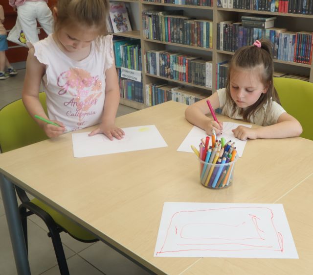 Na zdjęciu znajdują się dwie dziewczynki rysujące na białych kartkach przy stoliku, na środku którego stoją kredki. W tle znajdują się półki z książkami.