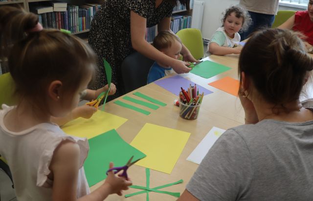 Na zdjęciu znajdują się dzieci wraz z rodzicami przy złączonych stołach, na których leżą kolorowe kartki i kredki.