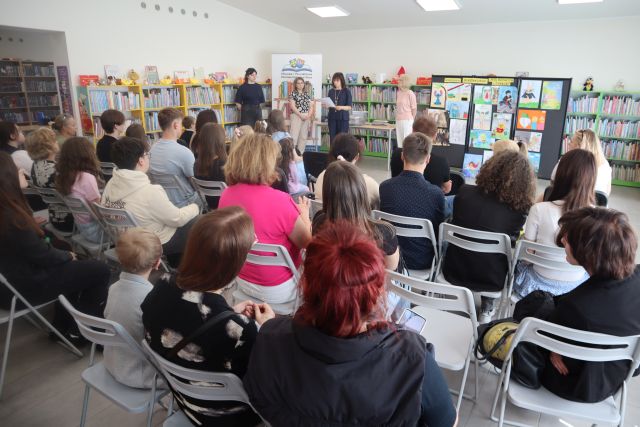 na krzesełkach siedzą laureaci konkursó i ich opiekunowie, w tle 3 bibliotekarki i dyrektorka biblioteki, regały z książkami, baner z nazwą i logo biblioteki oraz wystawa prac plastycznych