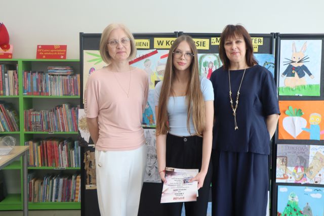 na zdjęciu bibliotekarka, dziewczyna z nagrodą książkową i dyplomem oraz dyrektorka biblioteki, w tle wystawa prac plastycznych