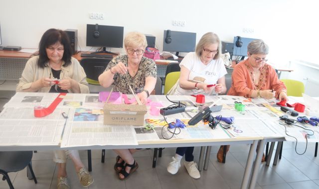Na zdjęciu znajdują się cztery kobiety siedzące przy złączonych stołach i robią kwiaty za pomocą wstążki, na stole przed nimi leżą gazety, wstążki, kleje na gorąco. Warsztaty odbywają się w sali komputerowej.