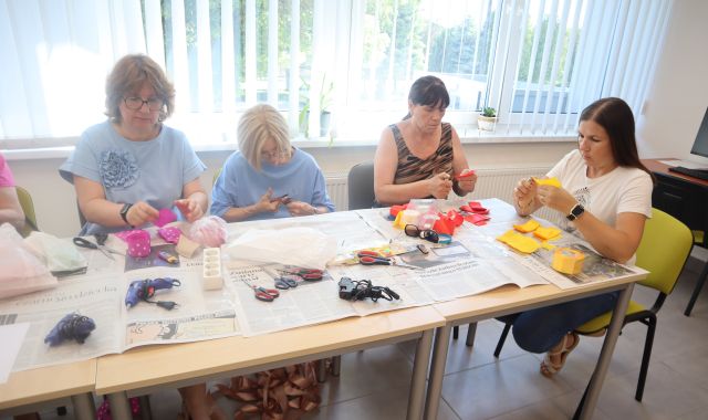 Na zdjęciu znajdują się cztery kobiety przy złączonych stołach i tworzą z kolorowych wstążek kwiaty. Na stolikach leżą gazety, kleje na gorąco i wstążki. Warsztaty odbywają się w sali komputerowej.