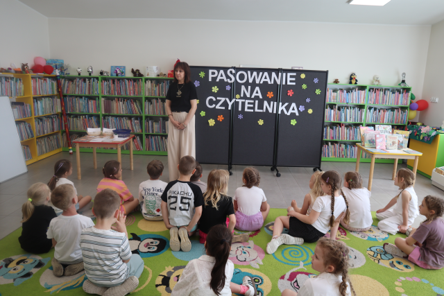 Na zdjęciu znajdują się przedszkolaki siedzące na dywanie, przed nimi stoi kobieta, za nią znajduje się czarna tablica z napisem Pasowanie na czytelnika oraz papierowe kwiaty, stoliki po lewej i prawej stronie oraz półki z książkami.