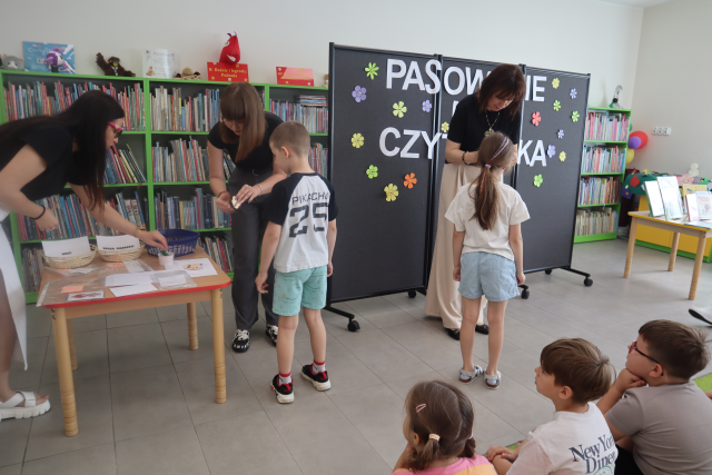 Na zdjęciu znajdują się trzy kobiety, które przypinają dzieciom kartki z imionami. Jedna z kobiet sięga kartkę ze stolika. W prawym dolnym rogu zdjęcia została uchwycona trójka dzieci. W tle znajduje się czarna tablica z napisem Pasowanie na Czytelnika oraz półki z książkami.