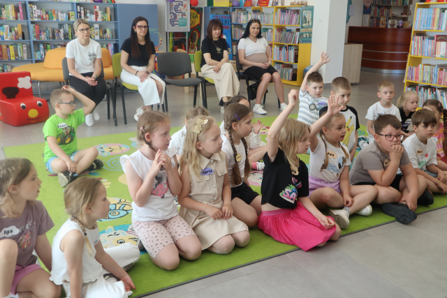 Na zdjęciu znajduje się grupa dzieci na dywanie i część z nich podnosi rękę do góry, w tle na krzesłach siedzą cztery kobiety. W tle znajdują się półki z książkami.