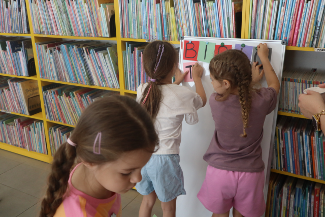 Na zdjęciu znajdują się dwie dziewczynki, które przypinają napis Biblioteka do białej magnetycznej tablicy, na zdjęciu znajduje się również dziewczynka, która przechodzi za dziewczynkami. W tle znajdują się półki z książkami.