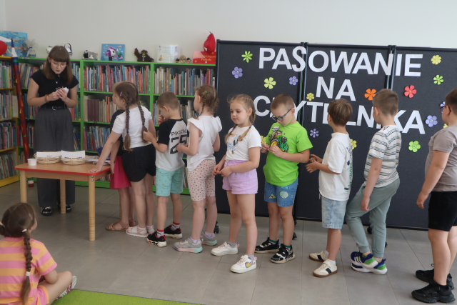 Na zdjęciu znajduje się kolejka dziecięciu dzieci, które stoją do stojącej po lewej stronie kobiety przy stoliku i czyta zagadki z kartki. W tle znajduje się czarna tablica z napisem Pasowanie na Czytelnika oraz półki z książkami.
