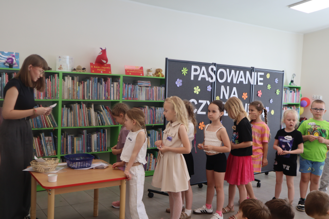 Na zdjęciu po lewej stronie znajduje się kobieta czytająca z kartki, przed nią stoi stolik z kartkami oraz koszyczkami, za stolikiem stoi kolejka przedszkolaków. W tle znajduje się czarna tablica z napisem Pasowanie na Czytelnika oraz półki z książkami.
