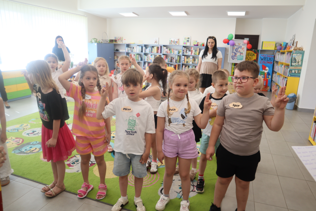 Na zdjęciu znajduje się grupa dzieci stojąca na dywanie, mają podniesione do góry dwa palce, za nimi stoją dwie kobiety. W tle znajdują się półki z grami oraz okna po lewej stronie.