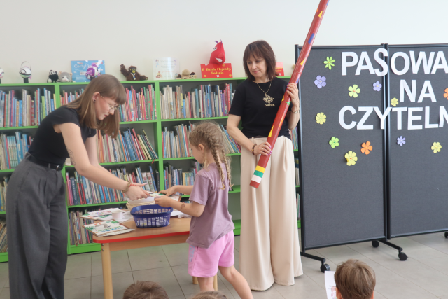 Na zdjęciu znajduje się dziewczynka, która dostaje od kobiety po lewej książkę, obok dziewczynki po prawej stoi kobieta z ołówkiem, obok niej stoi czarna tablica z napisem. W tle znajdują się półki z książkami.