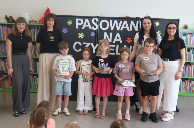 Na zdjęciu znajdują się cztery kobiety stojące razem do zdjęcia z piątką dzieci, które mają książki i dyplomy. Za nimi znajduje się tablica z napisem Pasowanie na czytelnika i półki z książkami w tle.
