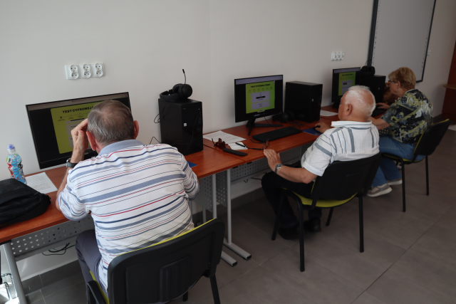 Na zdjęciu znajdują się trzy osoby przy komputerach i robią test. Warsztaty odbywają się w sali komputerowej.