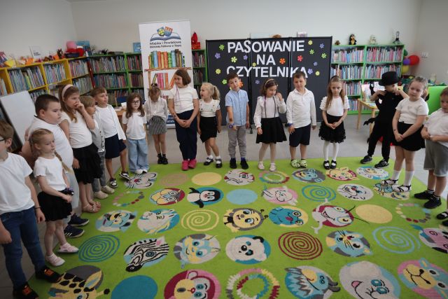 Na zdjęciu znajduje sie grupa dzieci stojąca wokół dywanu. Za nimi znajduje się tablica z napisem Pasowanie na Czytelnika, baner z logiem biblioteki i książkami. W tle znajdują się półki z książkami.