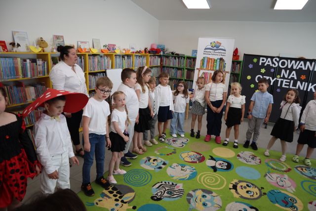 Na zdjęciu znajdują się przedszkolaki stojące wokół dywanu, chłopiec po lewej ma na głowie czapkę muchomora, za nimi stoi kobieta. W tle znajdują się półki z książkami, czarna tablica z napisem oraz baner z książkami i logiem biblioteki.