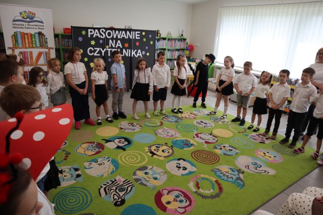Na zdjęciu stoją przedszkolaki dookoła zielonego dywanu. W tle znajdują się półki z książkami, tablica z napisem oraz okna.