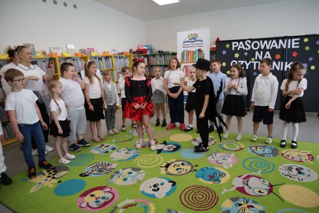 Na zdjęciu znajdują się dzieci wokół dywanu, na środku na dywanie stoi chłopczyk przebrany za żuka oraz dziewczynka ubrana za biedronkę. W tle znajdują się półki z książkami, czarna tablica z napisem oraz baner z książkami i logiem biblioteki.