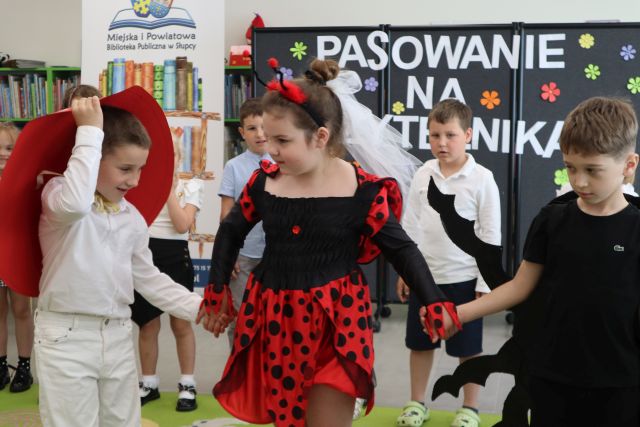 Na zdjęciu znajduje się trójka dzieci i trzymają się za ręce, dziewczynka ubrana za biedronkę, chłopiec po prawej za żuka i chłopiec po lewej za muchomora. W tle znajdują się stojące dzieci, czarna tablica z napisami i baner z logiem biblioteki i książkami.
