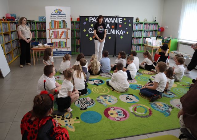 Na zdjęciu znajdują się elegancko ubrane dzieci, przed nimi stoi kobieta. W tle znajduje się czarna tablica z napisem, baner z logiem biblioteki i książkami, stolik po lewej stronie przy którym stoi kobieta oraz półki z książkami.