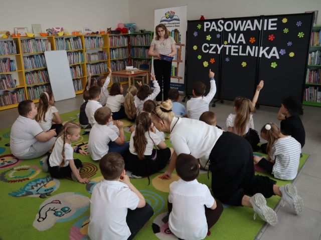 Na zdjęciu znajdują się dzieci na dywanie, część z nich ma podniesioną do góry rękę, pomiędzy nimi siedzi kobieta. Przed dywanem stoi kobieta. W tle znajduje się czarna tablica z napisem, baner z książkami i logiem biblioteki, półki z książkami oraz biała tablica po lewej stronie.