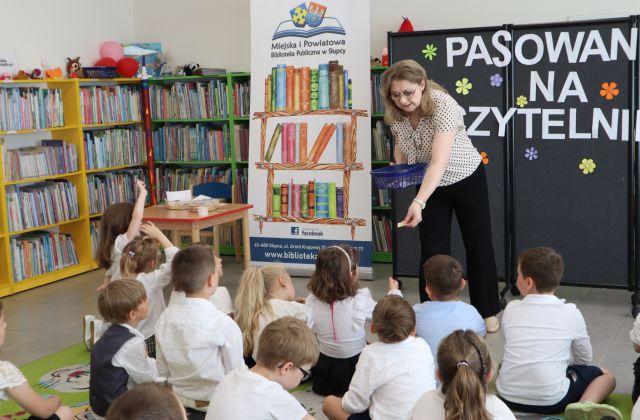 Na zdjęciu znajdują się siedzące na dywanie dzieci, przed nimi nachyla się kobieta z koszyczkiem i wręcza jednemu z nich papierowe słoneczko. W tle znajduje się czarna tablica z napisem, baner z książkami i logiem biblioteki, półki z książkami.