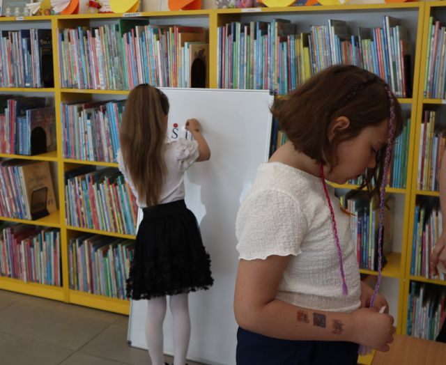 Na zdjęciu znajdują się dwie dziewczynki, jedna z nich przyczepia na białej tablicy literki, a druga przechodzi obok. W tle znajdują się półki z książkami.