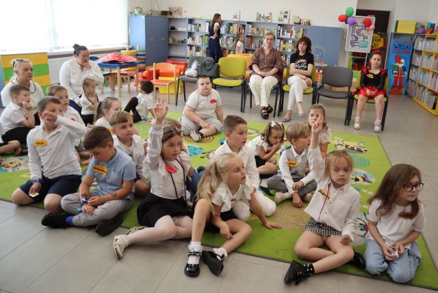Na zdjęciu znajdują się dzieci na dywanie, za nimi siedzą dwie kobiety na krzesłach. W tle znajdują się półki z grami.