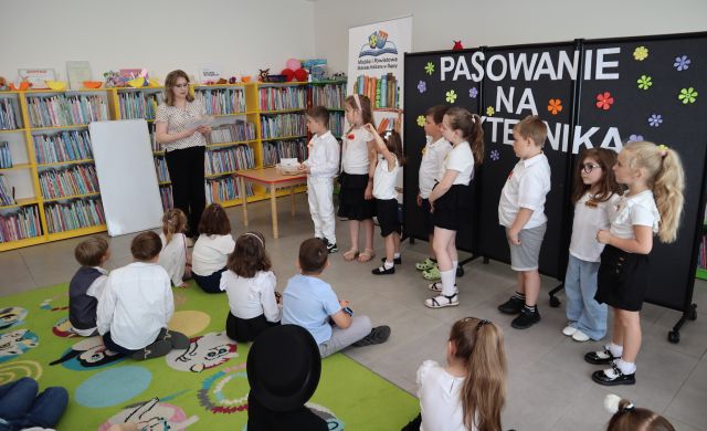 Na zdjęciu po lewej stronie siedzą dzieci na dywanie, a po prawej dzieci stoją w kolejce do zadania, przed nimi stoi kobieta. W tle znajduje się czarna tablica z napisem, baner z książkami i logiem biblioteki, półki z książkami oraz biała tablica po lewej stronie.
