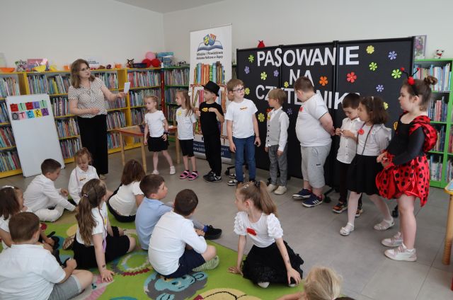 Na zdjęciu w lewym dolnym rogu znajdują się dzieci siedzące na dywanie, na przeciwko nich stoją dzieci, jeden za drugim, w kolejce do stojącej po lewo kobiety z kartką. W tle znajdują się półki z książkami, po prawej stronie znajduje się czarna tablica z napisem oraz baner z logiem biblioteki i książkami.