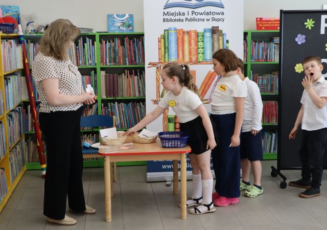 Na zdjęciu po prawej stronie znajdują się dzieci w kolejce do stolika, dziewczynka stojąca przy stoliku się nachyla i wrzuca karteczkę do koszyka, po drugiej stronie stołu stoi kobieta. W tle znajdują się półki z książkami, baner z książkami i logiem biblioteki oraz czarna tablica.