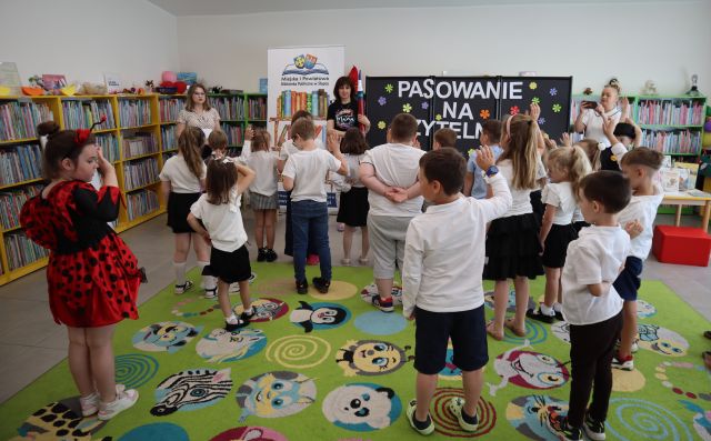 Na zdjęciu znajdują się dzieci stojące na dywanie, mają podniesione do góry dwa palce, przed nimi stoją dwie kobiety, jedna z nich ma duży ołówek. W tle znajdują się półki z książkami, czarna tablica z napisem oraz baner z logiem biblioteki i książkami.