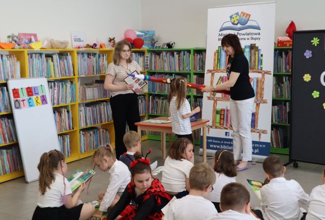 Na zdjęciu znajdują się dzieci na dywanie, przed nimi kobieta pasuje dziewczynkę dużym ołówkiem, po lewej stronie stoi kobieta z książką i dyplomem. W tle znajdują się półki z książkami, baner z logiem biblioteki i książkami oraz czarna tablica po prawej stronie, a po lewej biała tablica z kolorowym napisem.