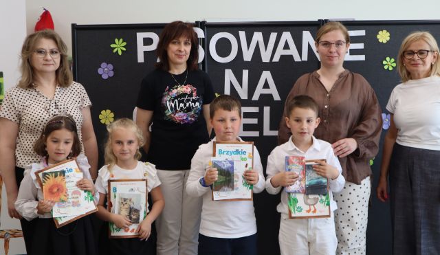 Na zdjęciu znajdują się cztery kobiety stojące do zdjęcia z czwórką dzieci, którzy trzymają książki z dyplomami oraz upominki. W tle znajduje się czarna tablica z białym napisem.