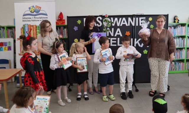Na zdjęciu znajdują się trzy kobiety, dwie z nich wraz z piątką dzieci stoją po lewej stronie, po prawej stoi kobieta z wyciągniętą ręką w której trzyma torbę z cukierkami. W tle znajduje się czarna tablica z napisem, baner z logiem biblioteki i książkami oraz półki z książkami.