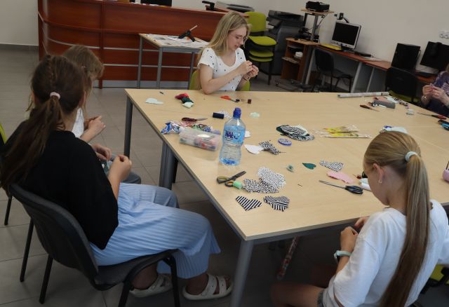 Na zdjęciu znajdują się dwie dziewczynki oraz kobieta przy złączonych stołach i szyją z kolorowych materiałów znajdujących się na stole wraz z nożyczkami, igłami, nitkami. Warsztaty odbywają się w sali komputerowej.