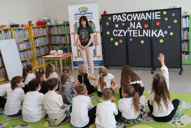 Na zdjęciu znajdują się dzieci na dywanie, a przed nimi stoi kobieta. W tle znajduje się czarna tablica z napisem Pasowanie na Czytelnika, baner z logiem biblioteki i książkami, półki z książkami i po lewej stronie oparta na nich biała tablica.
