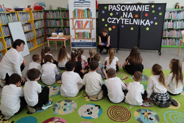 Na zdjęciu znajdują się przedszkolaki na dywanie, przed nimi kuca kobieta z plastikowym koszyczkiem, W tle znajduje się czarna tablica z napisem Pasowanie na Czytelnika, baner z logiem biblioteki i książkami, stolik, półki z książkami i po lewej stronie oparta na nich biała tablica.