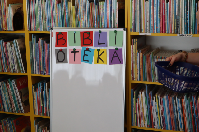 Na zdjęciu znajduje się biała tablica z przyczepionym kolorowym napisem biblioteka, tablica oparta jest o półki z książkami.