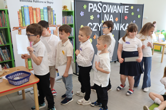 Na zdjęciu znajdują się stojące w kolejce do stolika po lewej dzieci. W tle znajduje się czarna tablica z napisem Pasowanie na Czytelnika, baner z logiem biblioteki i książkami, półki z książkami.