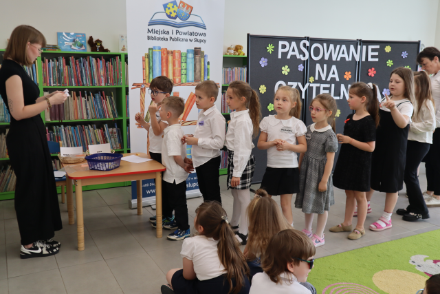 Na zdjęciu po lewej stronie stoi kobieta i czyta zagadki z kartki, przed nią stoi stolik z kartkami, a za stolikiem stoją dzieci w kolejce. W tle znajdują sę półki z książkami, baner z książkami i logiem biblioteki i czarna tablica z napisem.