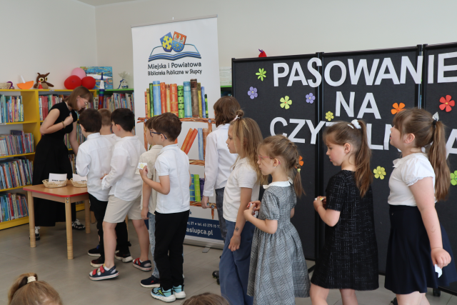 Na zdjęciu znajduje się kolejka dzieci do kobiety przy stoliku, która czyta zagadki. W tle znajduje się czarna tablica z napisem Pasowanie na Czytelnika, baner z logiem biblioteki i książkami, półki z książkami.
