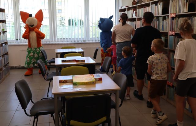 Na zdjęciu znajduje się maskotka lisa oraz niebieskiego kota, którzy prowadzą dzieci wraz z rodzicami w pociągu na środku stoją stoły, po lewej i prawej stronie znajdują się półki z książkami, a w tle okna.
