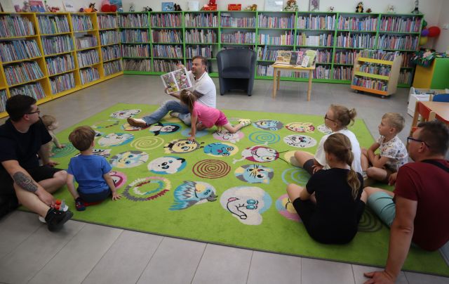 Na zdjęciu znajduje się trójka ojców wraz z piątką dzieci na dywanie, jeden z panów siedzący na środku czyta książkę. W tle znajduje się stolik z książkami, obok fotel oraz półki z książkami.